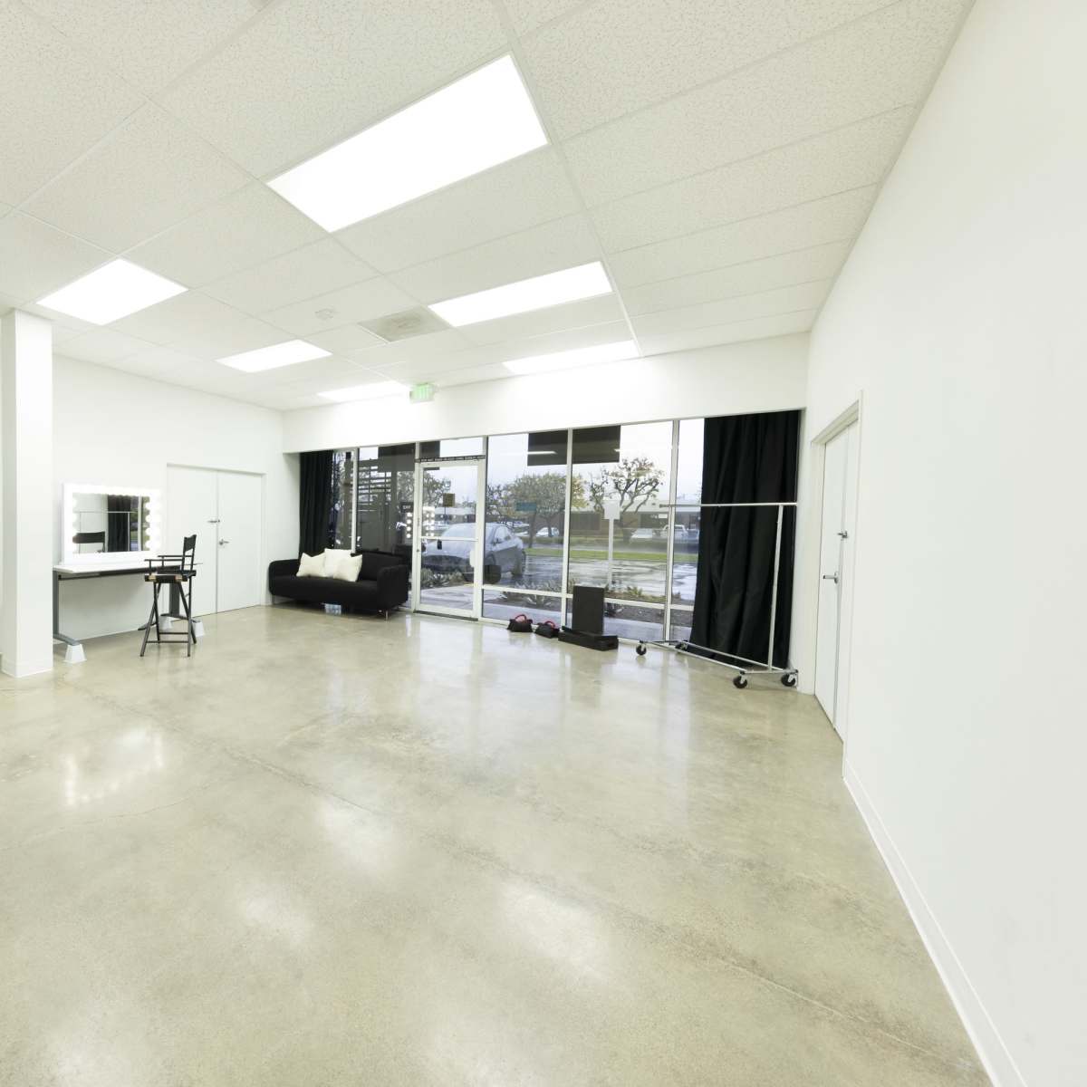 An empty room with polished concrete flooring, a large window, a black curtain, a mirror, a vanity setup, and a pink chair.