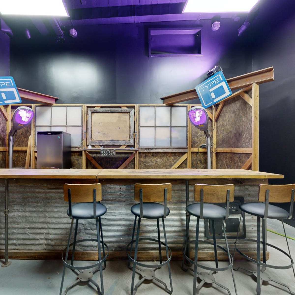 A rustic bar with a wooden countertop and metal stools, illuminated by purple lights and featuring a vintage-style wooden backdrop.