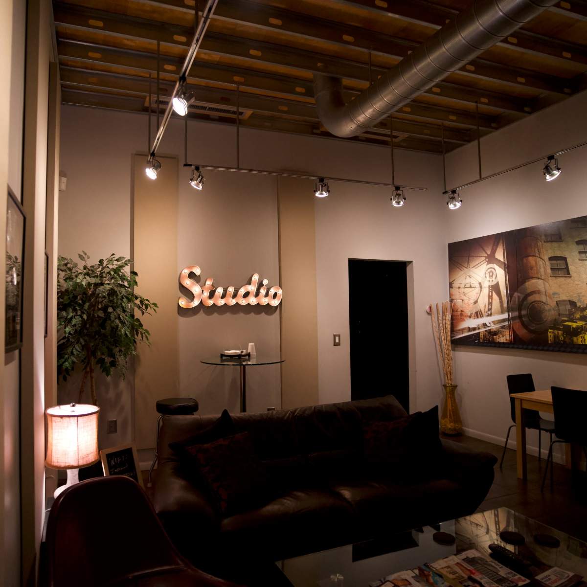 A modern studio space featuring a dark sofa, a round table, a decorative plant, and wall art, illuminated by ceiling lights and a lamp.