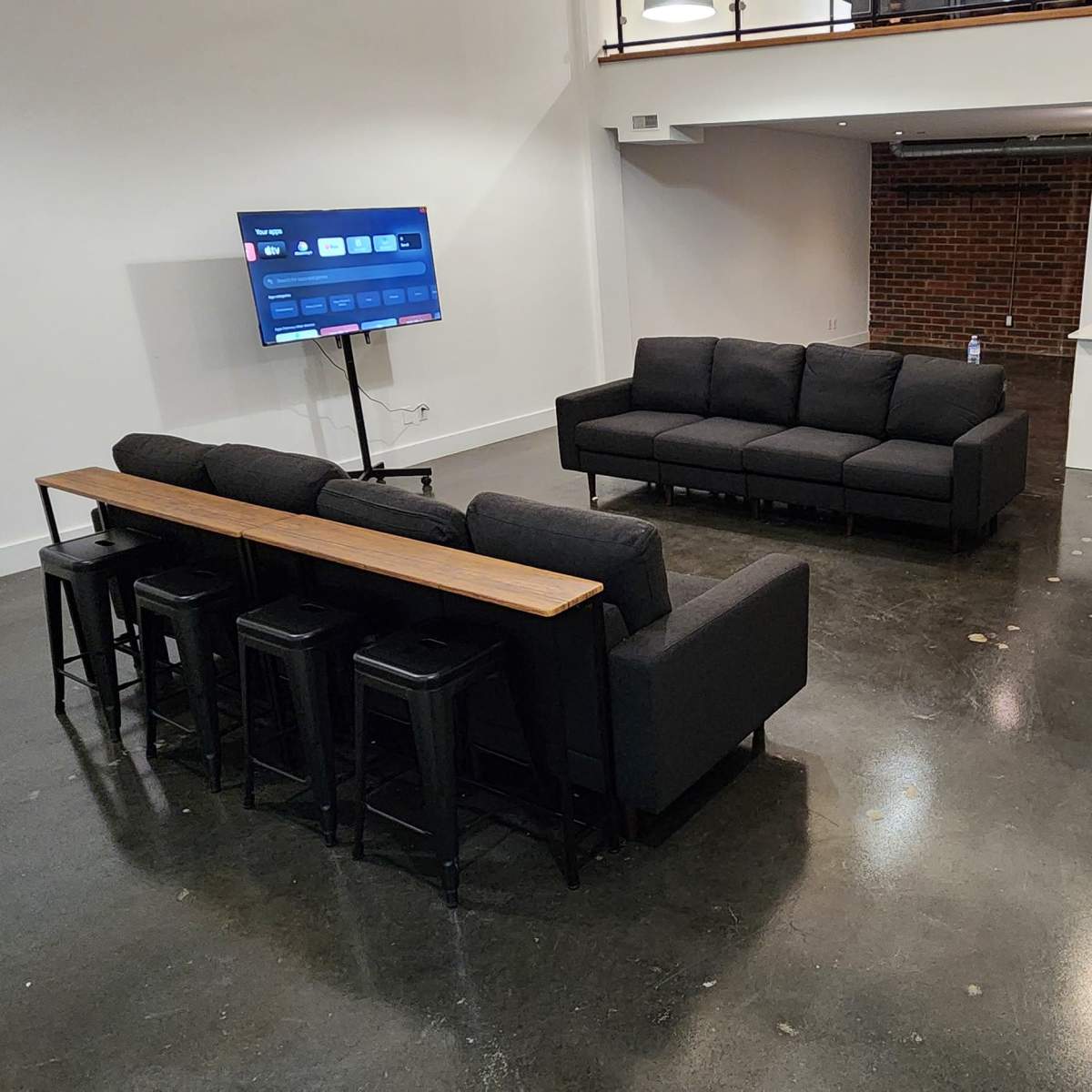 A spacious living area with a black sofa set facing a television, and a dining table with stools behind it, while a staircase leads to an upper level.