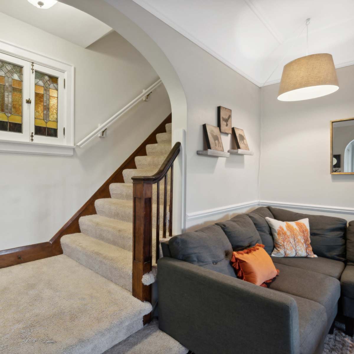 A staircase with a stained glass window leads from a carpeted landing to a living area that features a gray couch with orange accents.