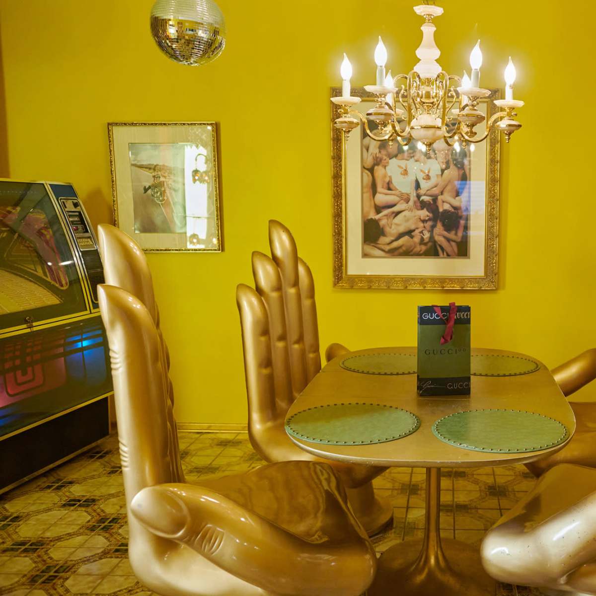 A round table with gold hand-shaped chairs is set in a brightly colored yellow dining room, featuring a chandelier and a framed artwork on the wall.