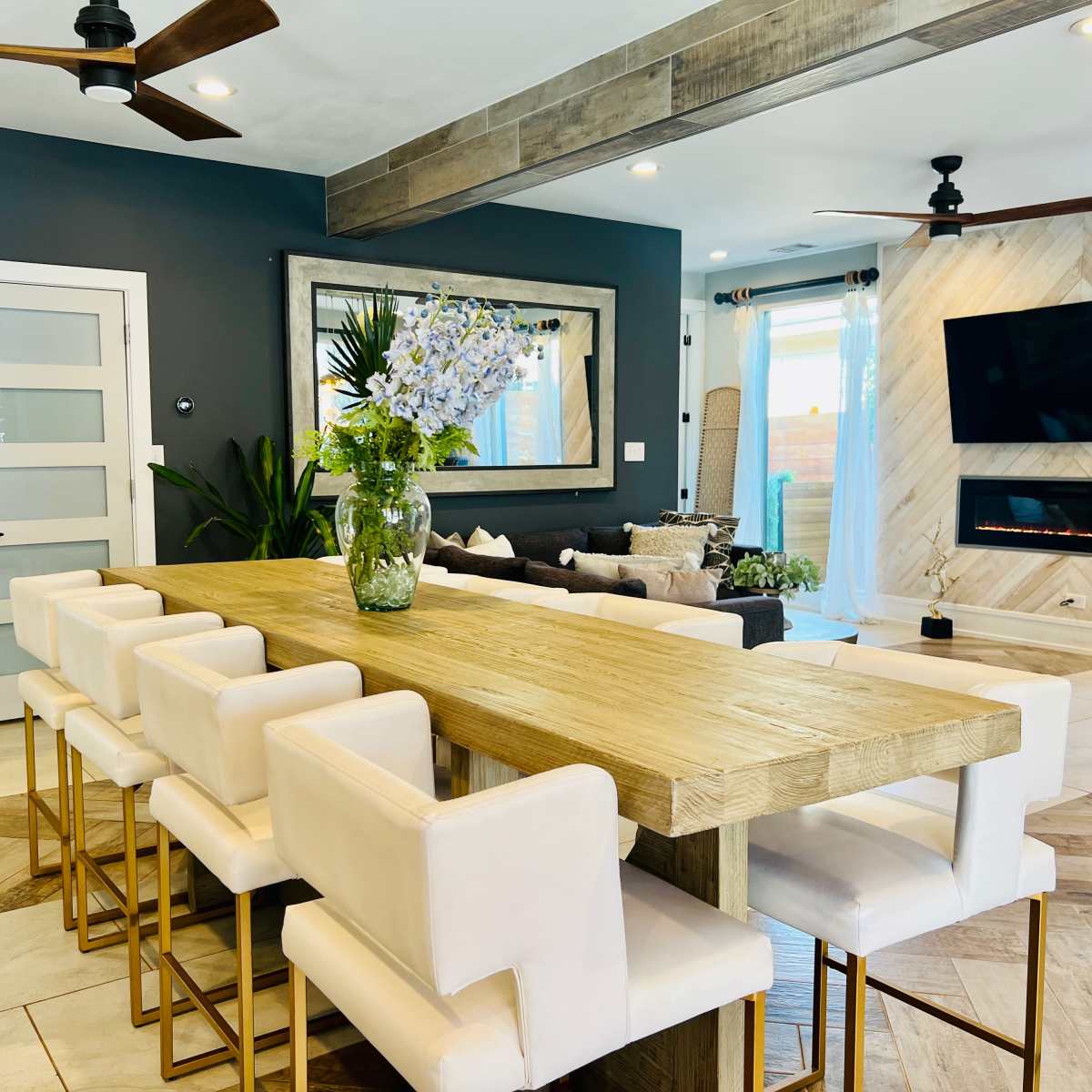 A modern dining area features a long wooden table surrounded by white bar stools, with a vase of flowers as a centerpiece and a wall-mounted television and fireplace in the background.