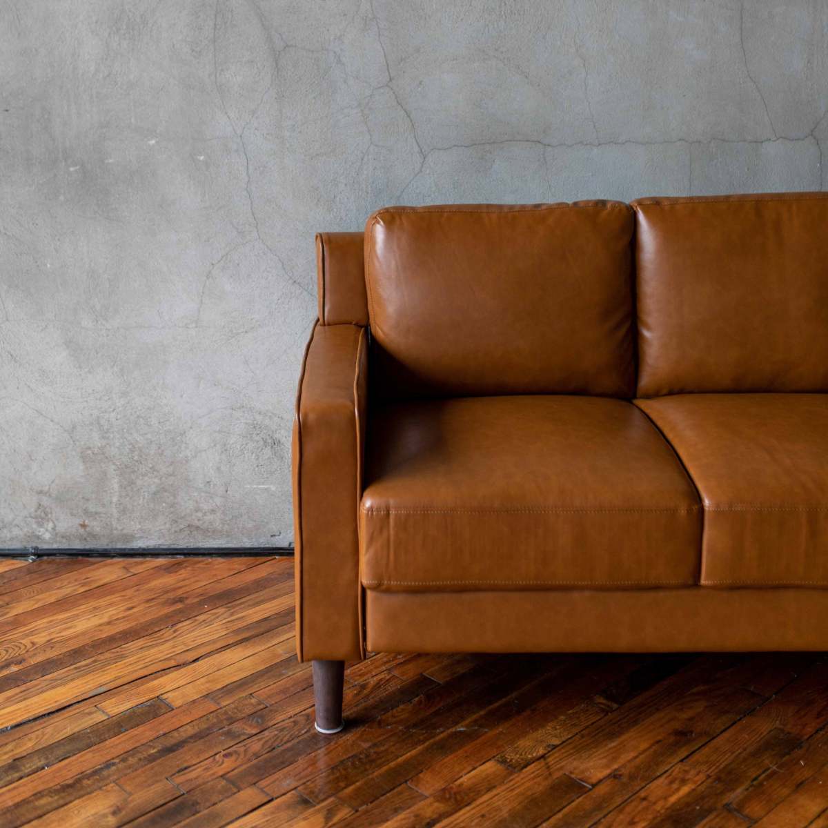 A brown leather sofa is positioned against a textured gray wall with visible cracks, on a wooden floor.