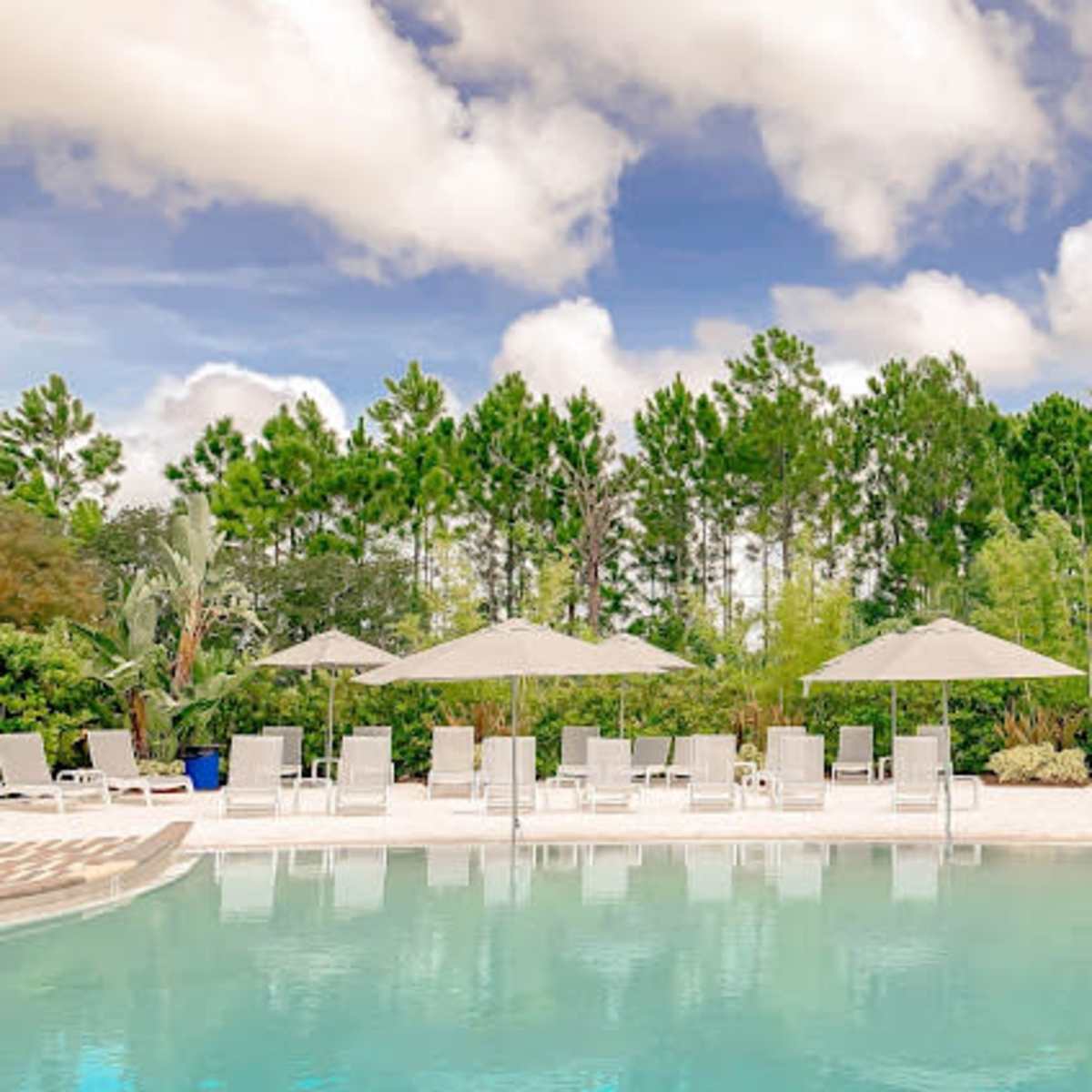 A clear swimming pool is surrounded by sun loungers and shaded umbrellas, with lush greenery in the background.