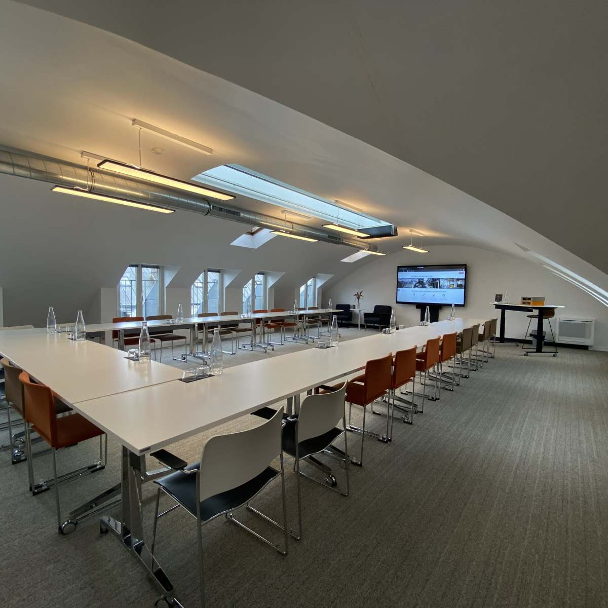 A spacious meeting room features a long table with chairs, a large screen, and skylights illuminating the area.