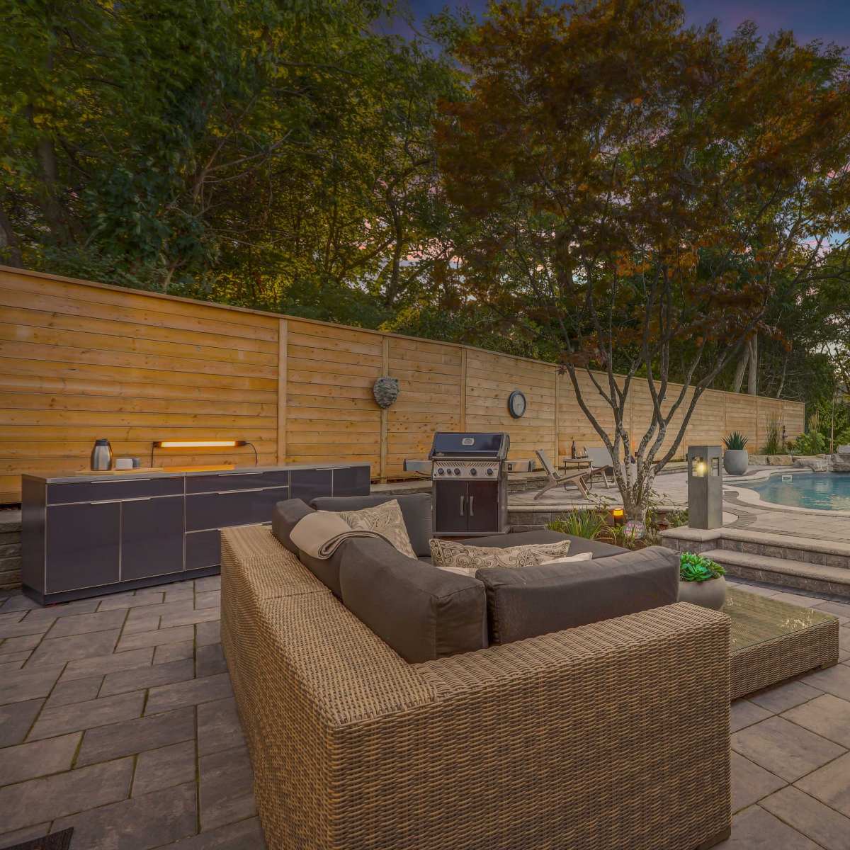 A backyard patio area with a stone path leading to a swimming pool, surrounded by trees and a wooden fence, featuring outdoor seating and a barbecue grill.