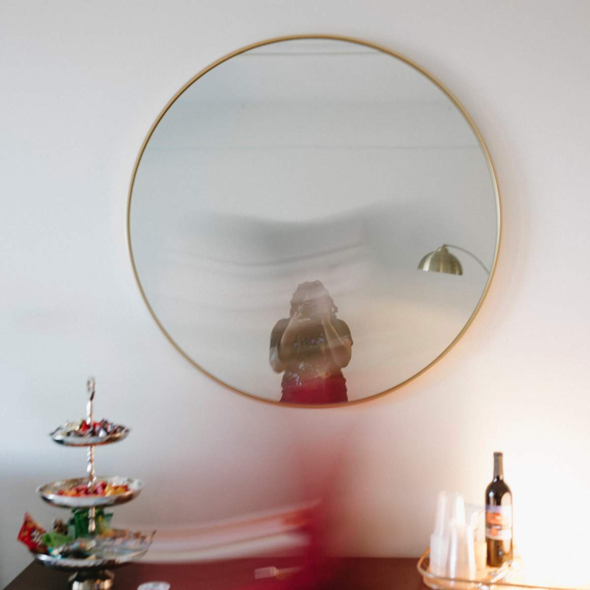 A round mirror reflects a blurred figure in a room with a wine bottle and a tiered stand of snacks.