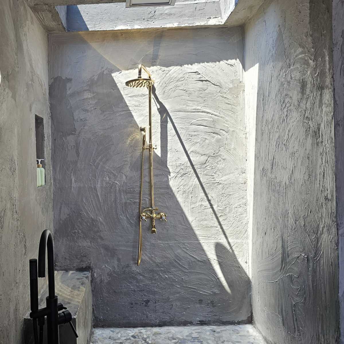 A modern shower area with textured gray walls, a gold shower fixture, and a hanging light bulb above a single rose.