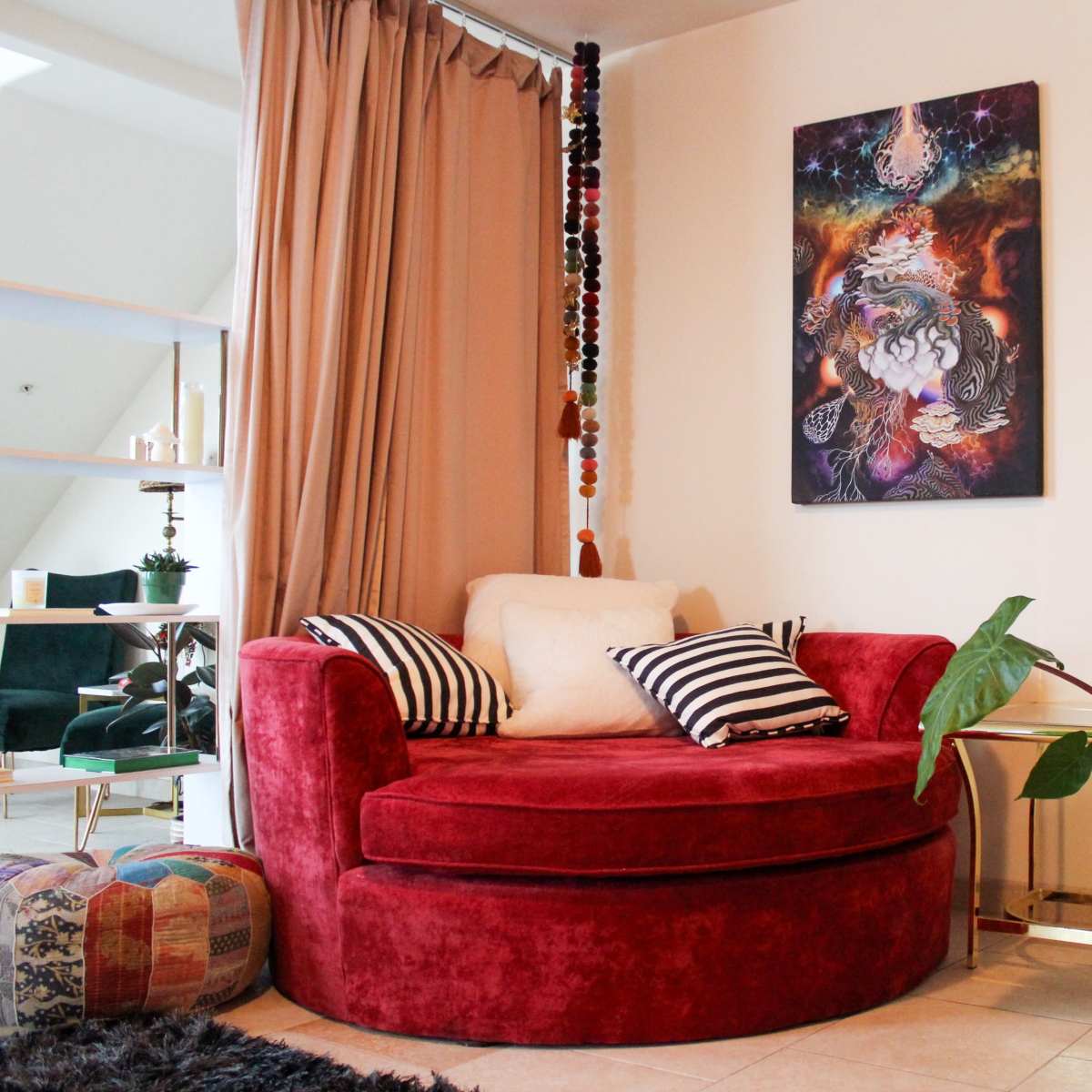 A cozy living space featuring a red circular sofa with pillows, a potted plant, and a colorful ottoman, all set against a wall adorned with a vibrant artwork.