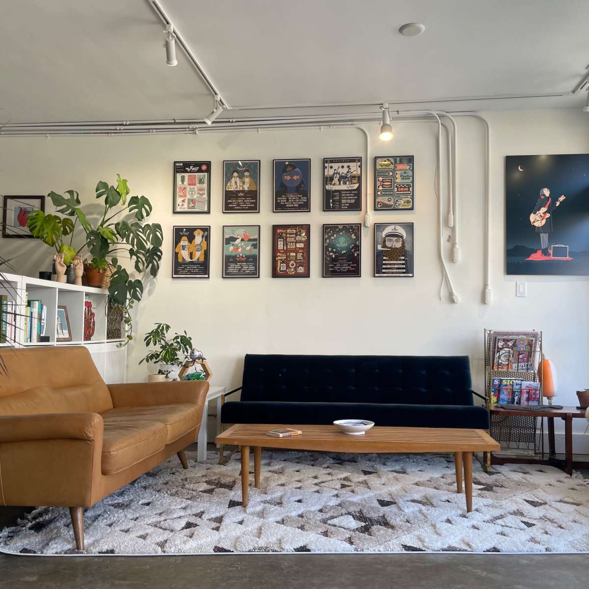 A modern living space featuring a dark blue sofa, a brown leather armchair, a wooden coffee table, and several framed posters on the wall, with plants and a decorative rug on the floor.