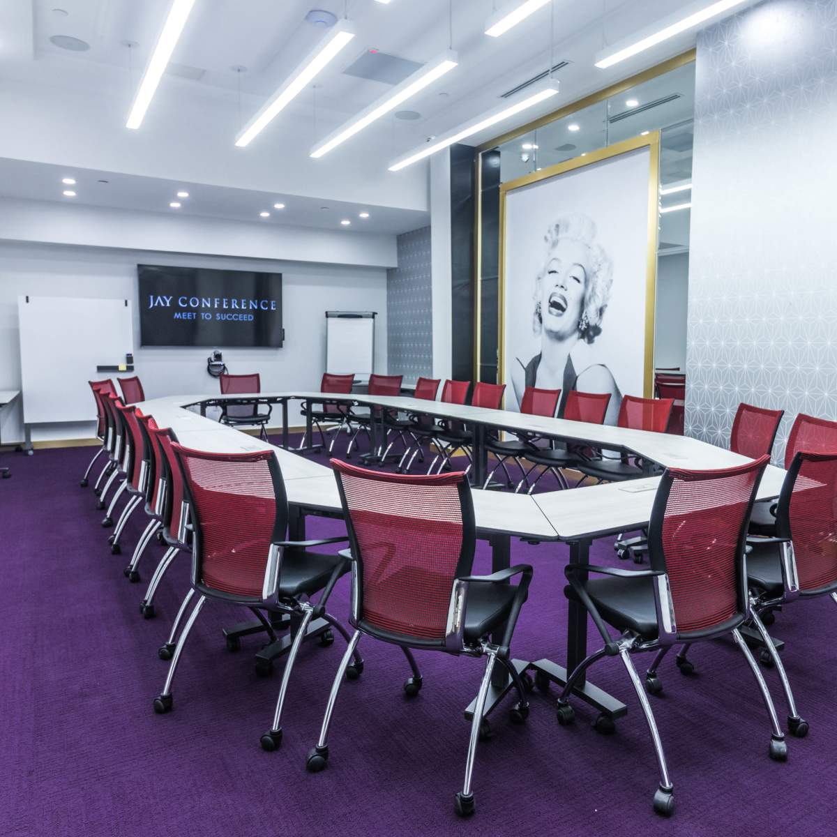 A modern conference room features a long, oval table surrounded by red mesh chairs, with a large image of a smiling woman on the wall.