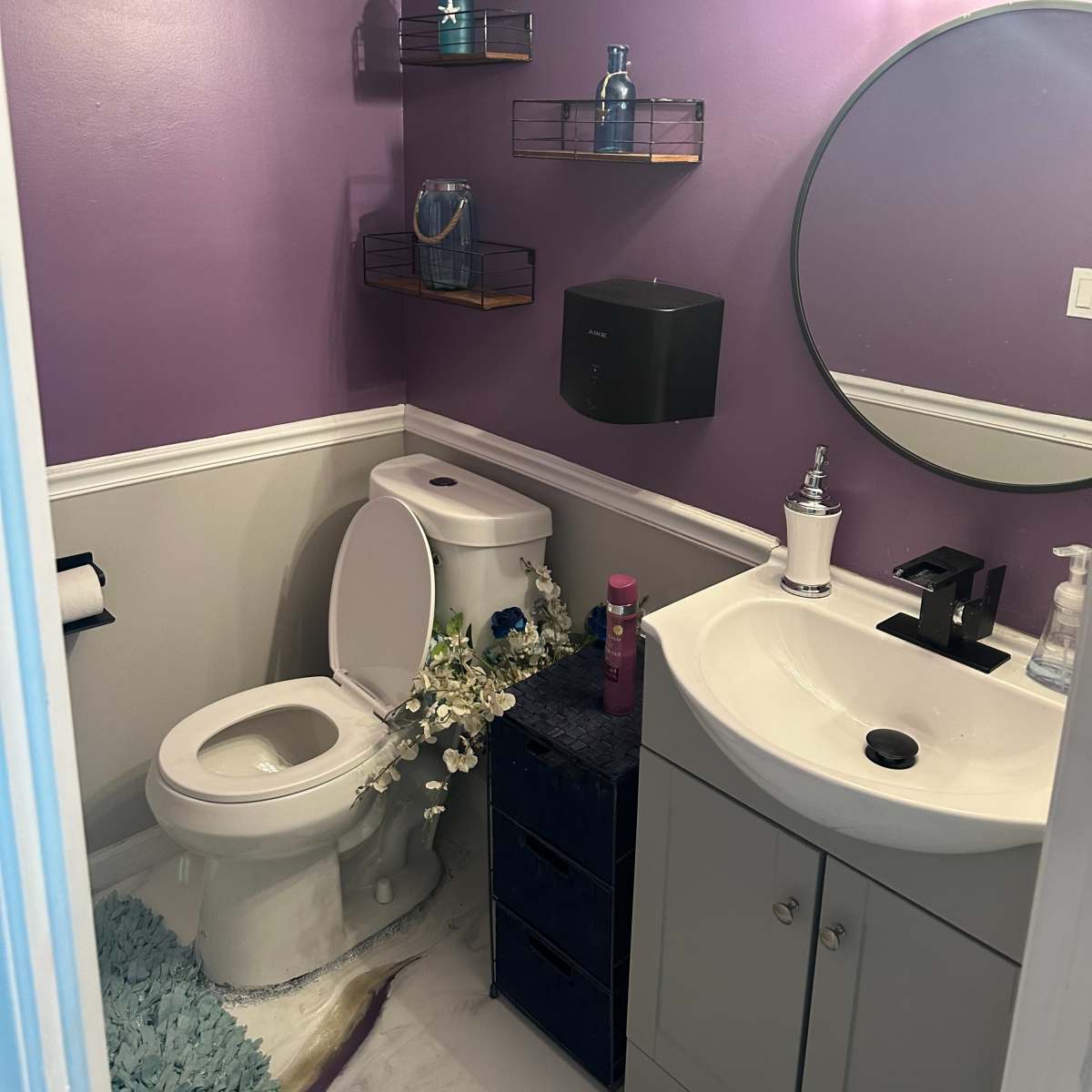 A small bathroom features a purple wall, a toilet, a sink with a dark cabinet, and decorative shelves with various toiletries.