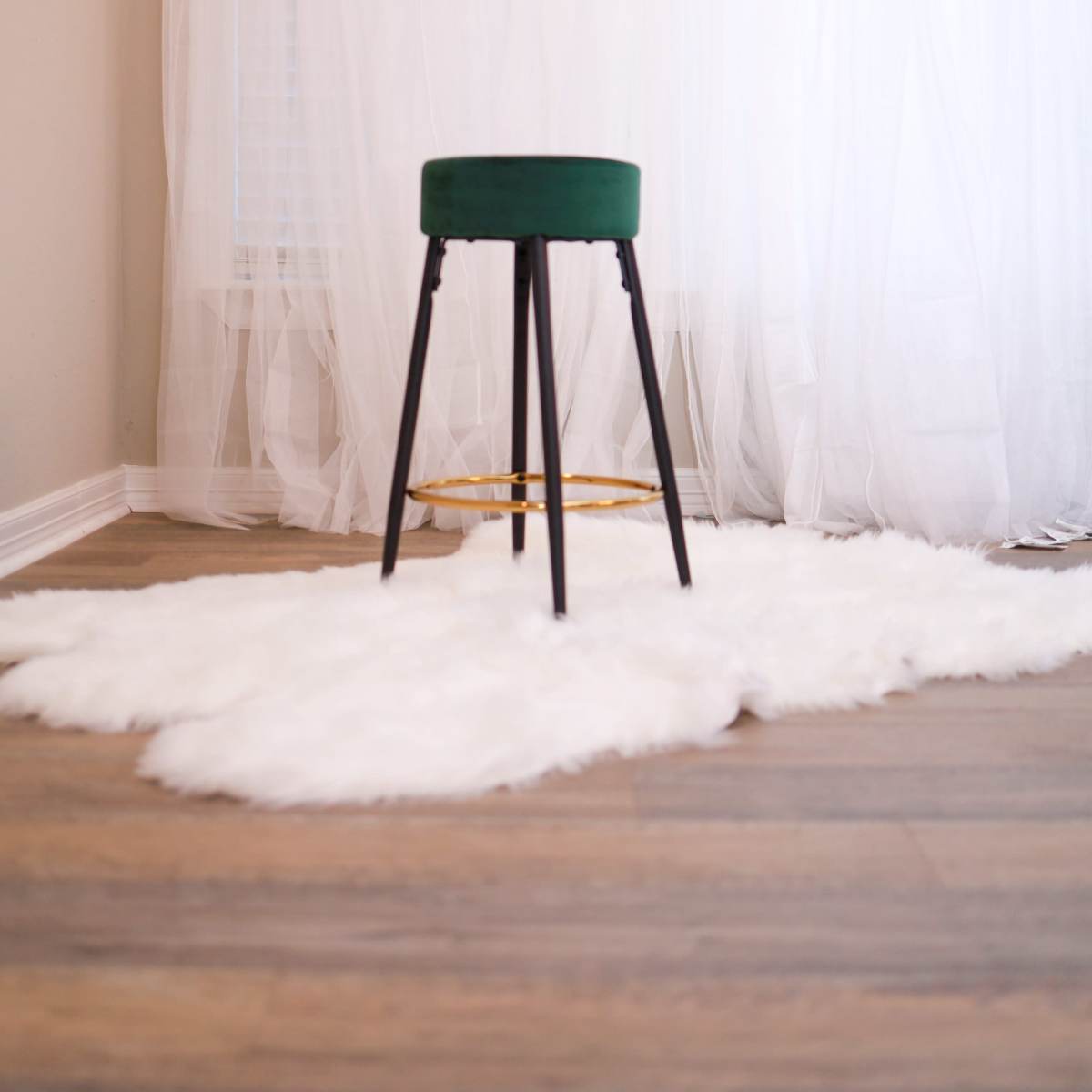 A green stool with black legs stands on a white faux fur rug in front of sheer white curtains.