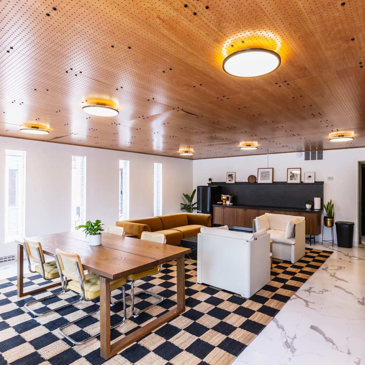 A modern interior space featuring a seating area with a brown couch, a dining table, and a sleek kitchen counter against a backdrop of a wooden ceiling and large windows.