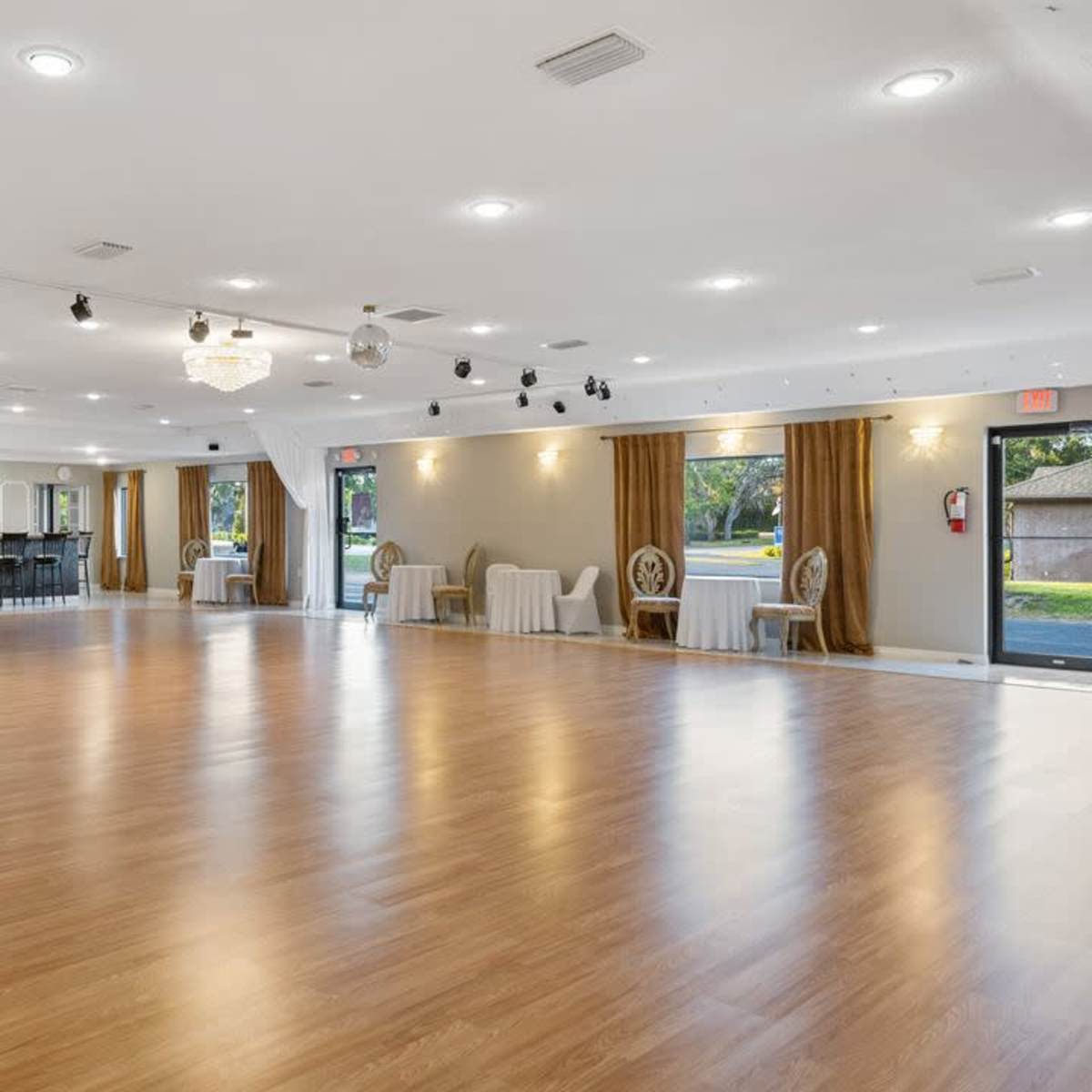 A spacious event hall with wooden flooring, large windows, and draped white curtains, ready for gatherings.