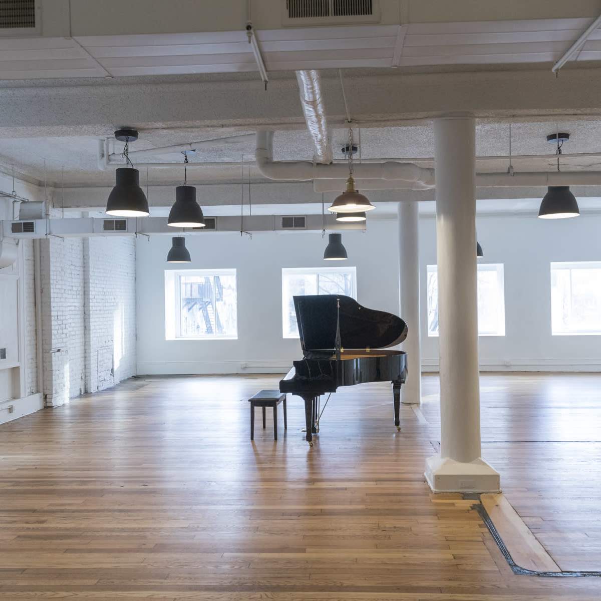 A grand piano sits alone in the center of a spacious, empty room with wooden floors and multiple windows.