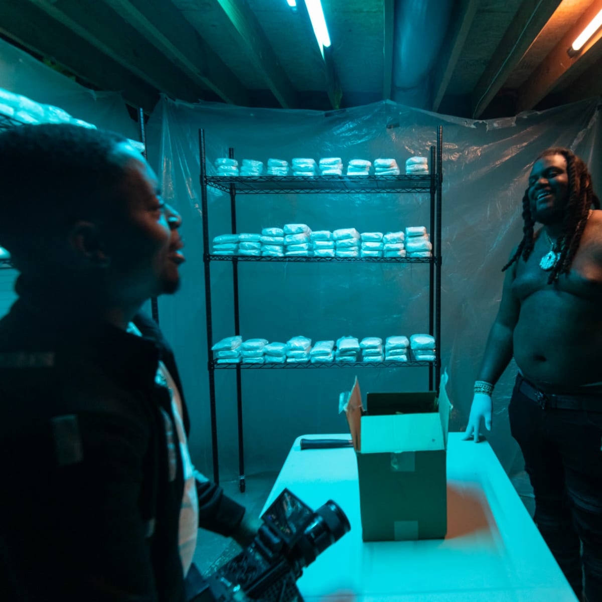 Two men stand in a dimly lit basement with blue lighting, beside a table and shelves lined with white packages.