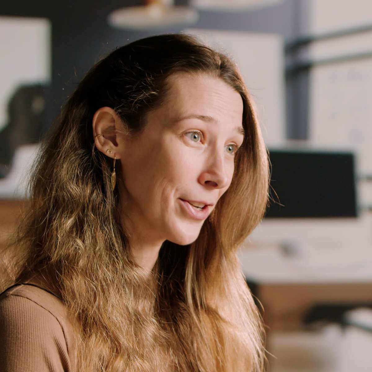 A woman with long, wavy hair is speaking in an office setting with furniture and greenery in the background.
