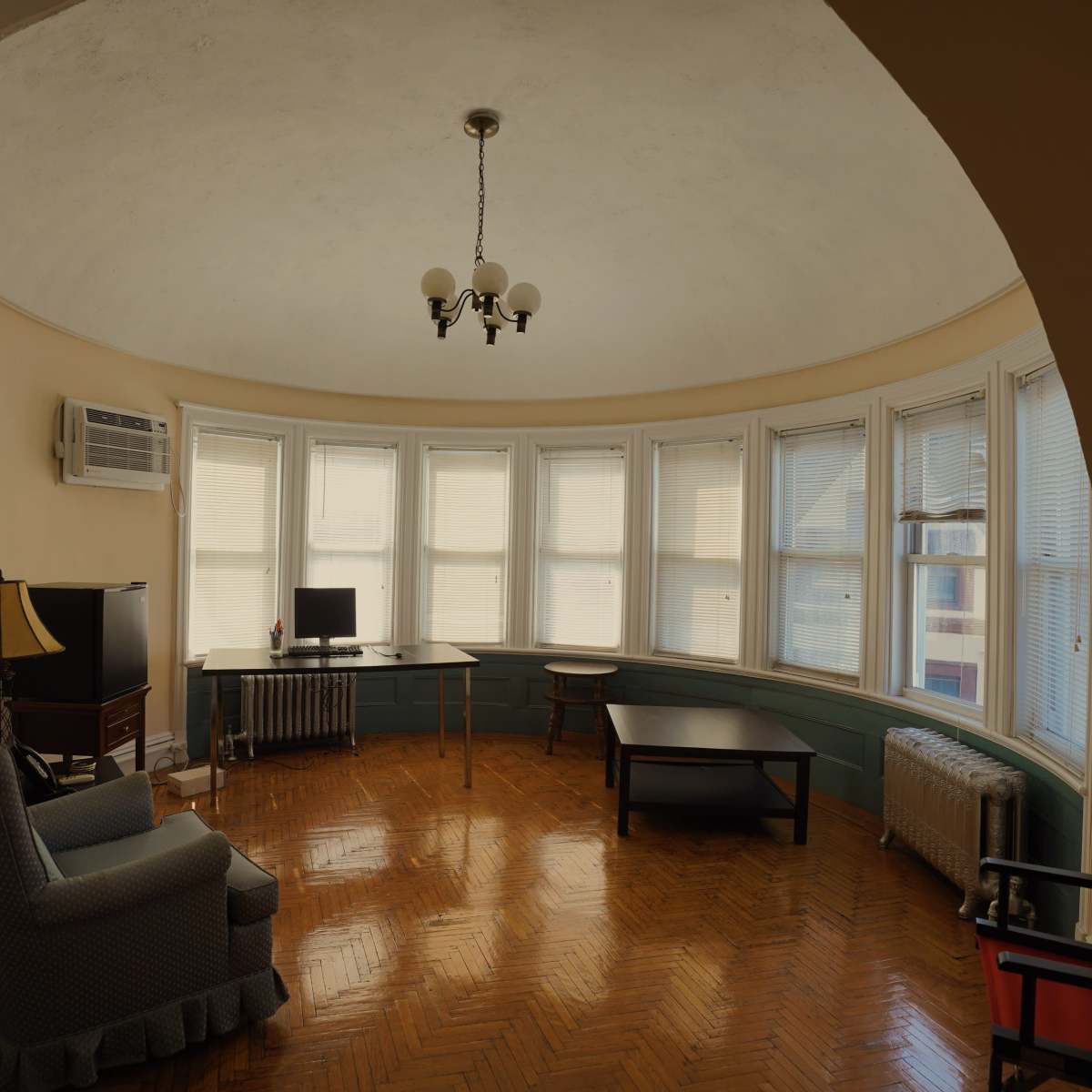 A curved room features a seating area, a desk with a computer, and several windows allowing light to enter.