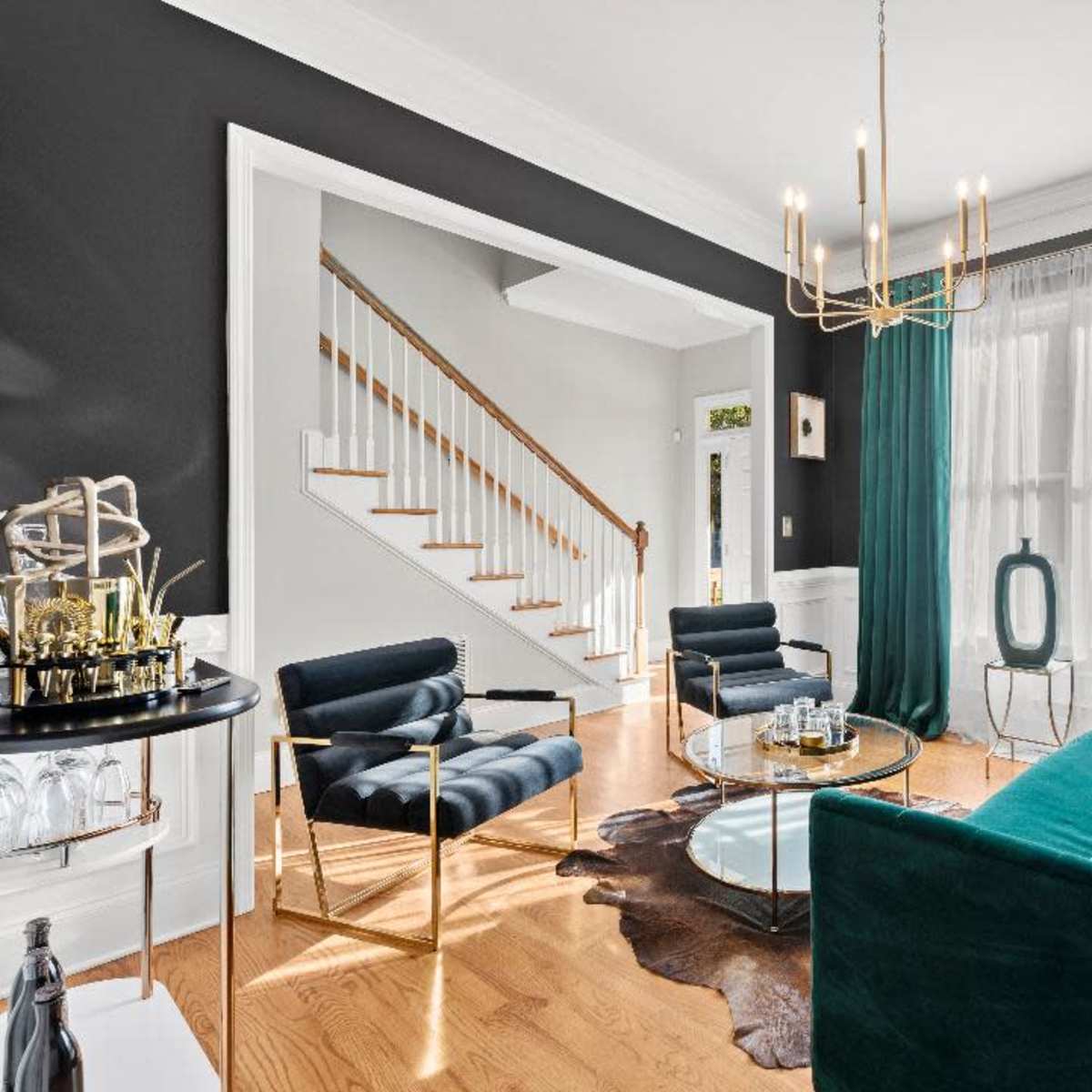 A modern living room featuring a green velvet sofa, black armchairs, a glass coffee table, and large windows dressed with sheer and dark curtains.