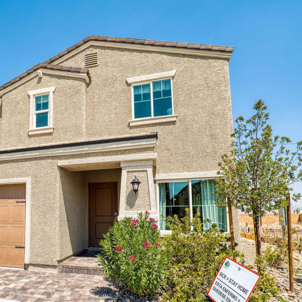 A two-story beige house with a brown garage door and large windows is situated on a paved driveway.