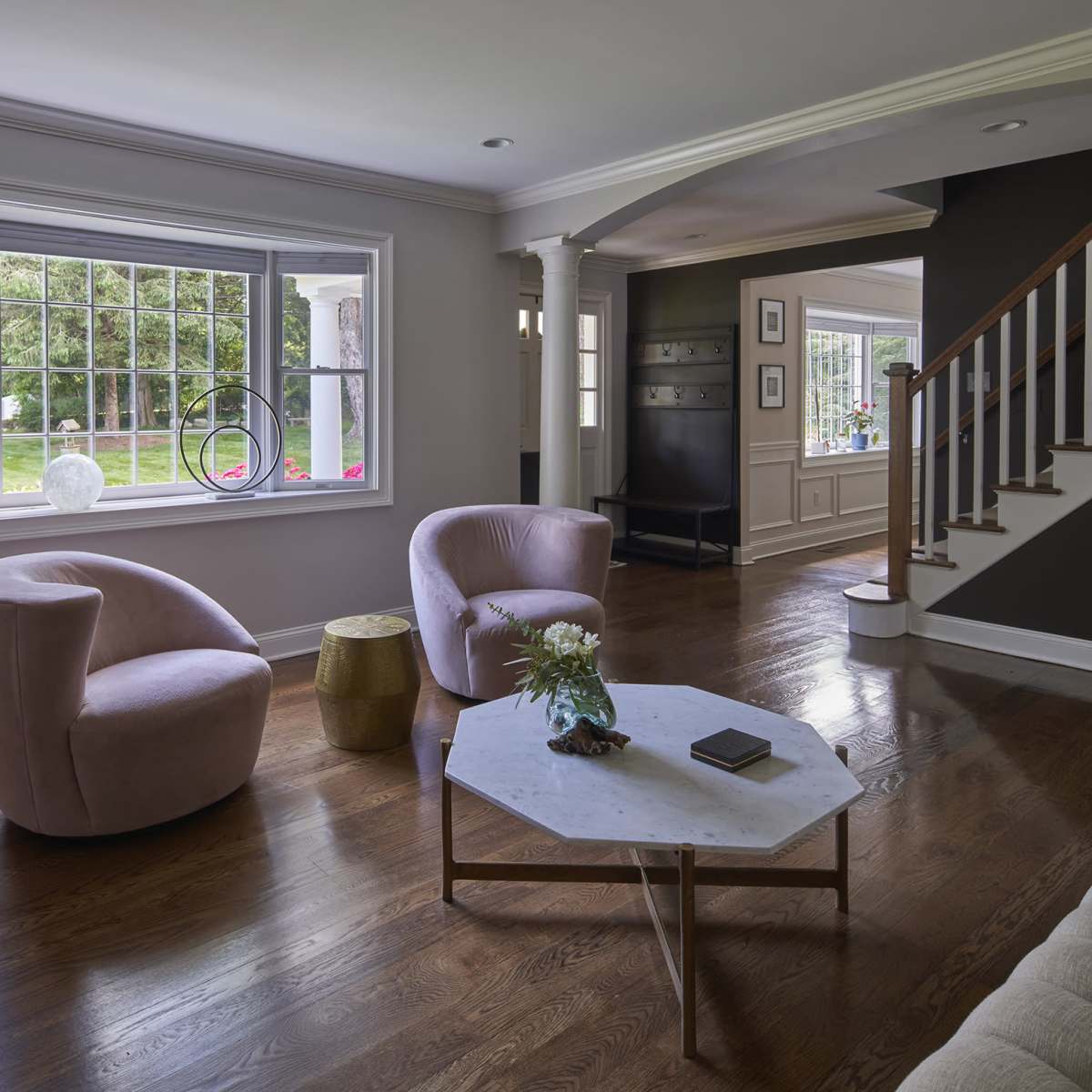 A light-filled living room with two pink chairs, a marble coffee table, and a staircase leading to another room.