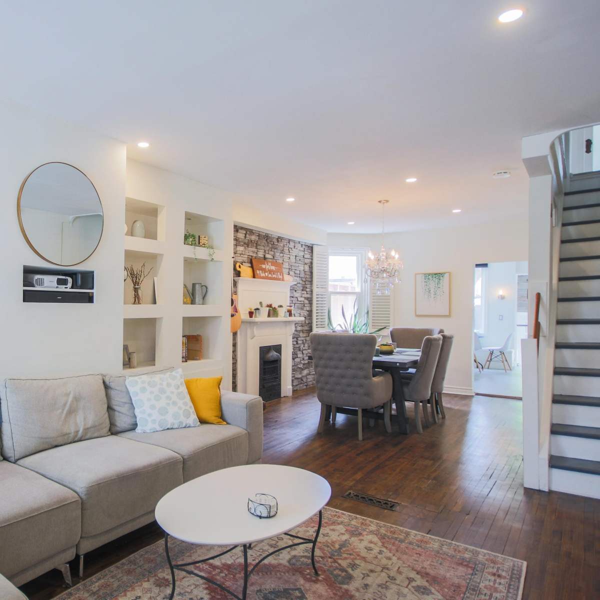 A modern living room with a gray sectional sofa, built-in shelves, a circular mirror, and a dining area in the background.