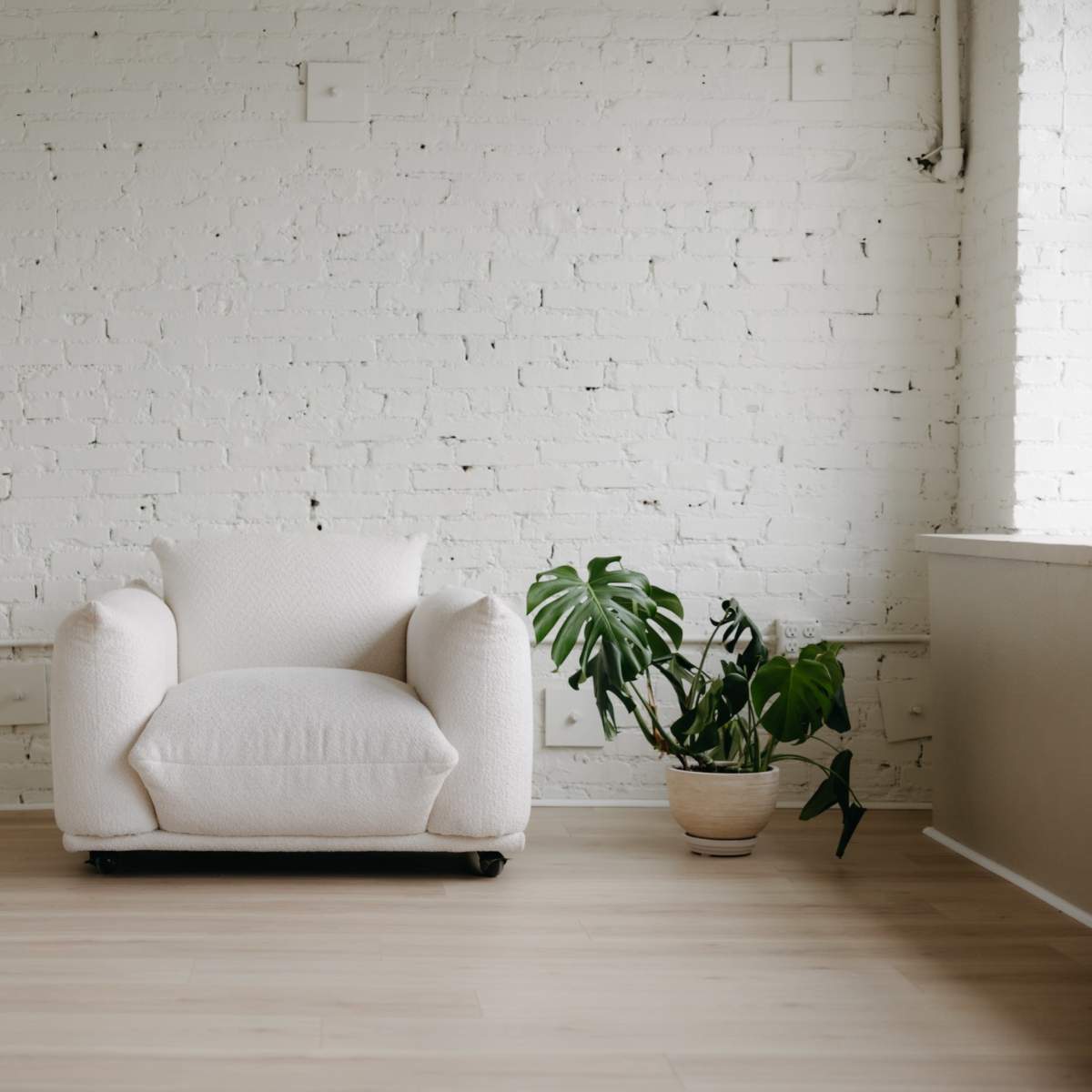 A single white armchair is positioned in a minimally furnished room with a potted plant and white brick walls.