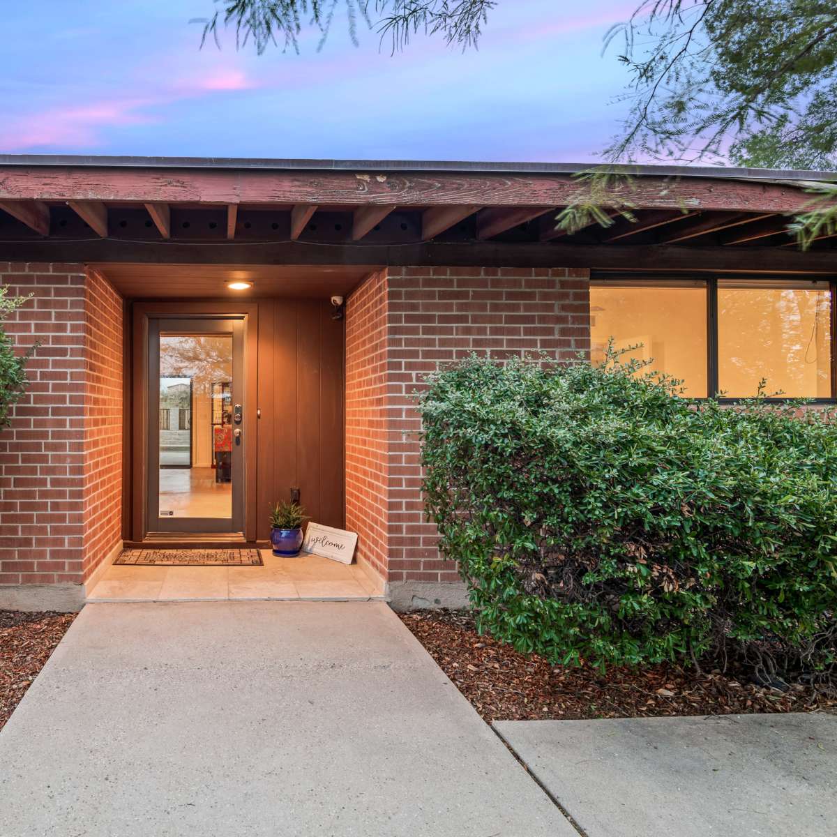 The entrance of a brick building framed by neatly trimmed hedges and a concrete pathway leading to the door.