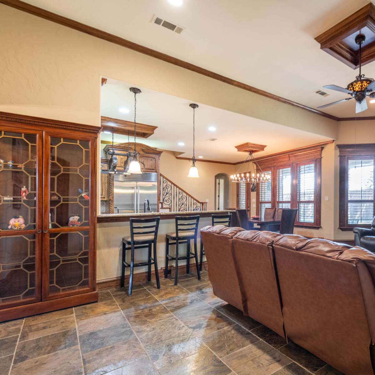 A spacious living room with a brown leather sofa, a stone fireplace, a wooden dining area, and large windows allowing natural light.