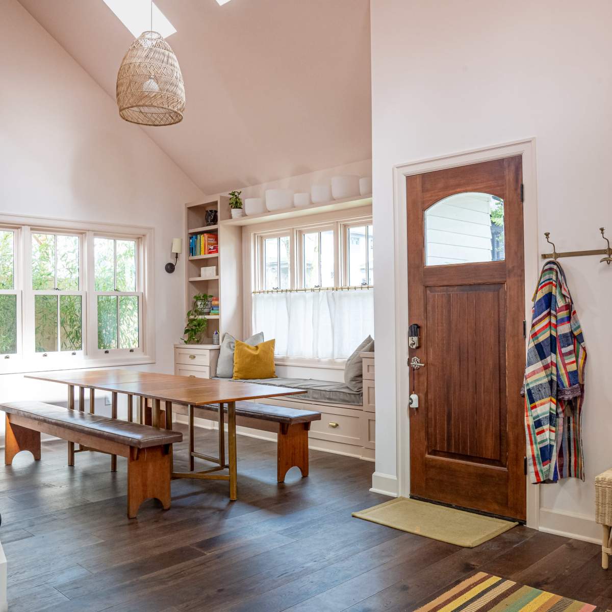 The interior of a home features a dining table and benches, a window seat with cushions, a wooden front door, and a staircase with a black railing.