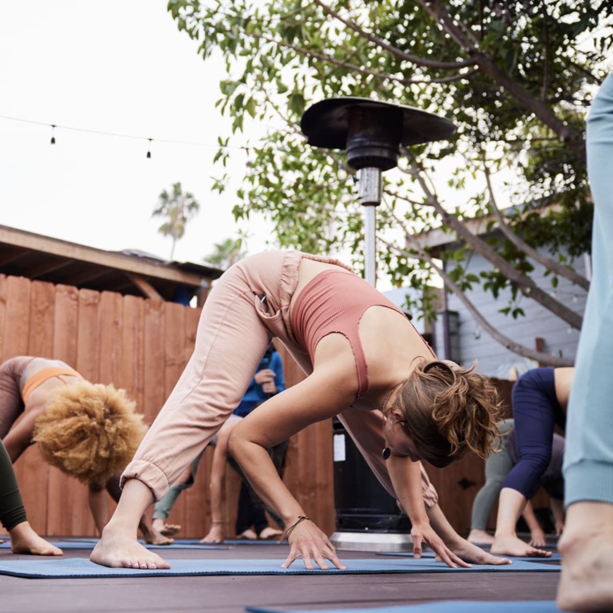 A group of people practices yoga outdoors on mats near a wooden fence and a heater.
