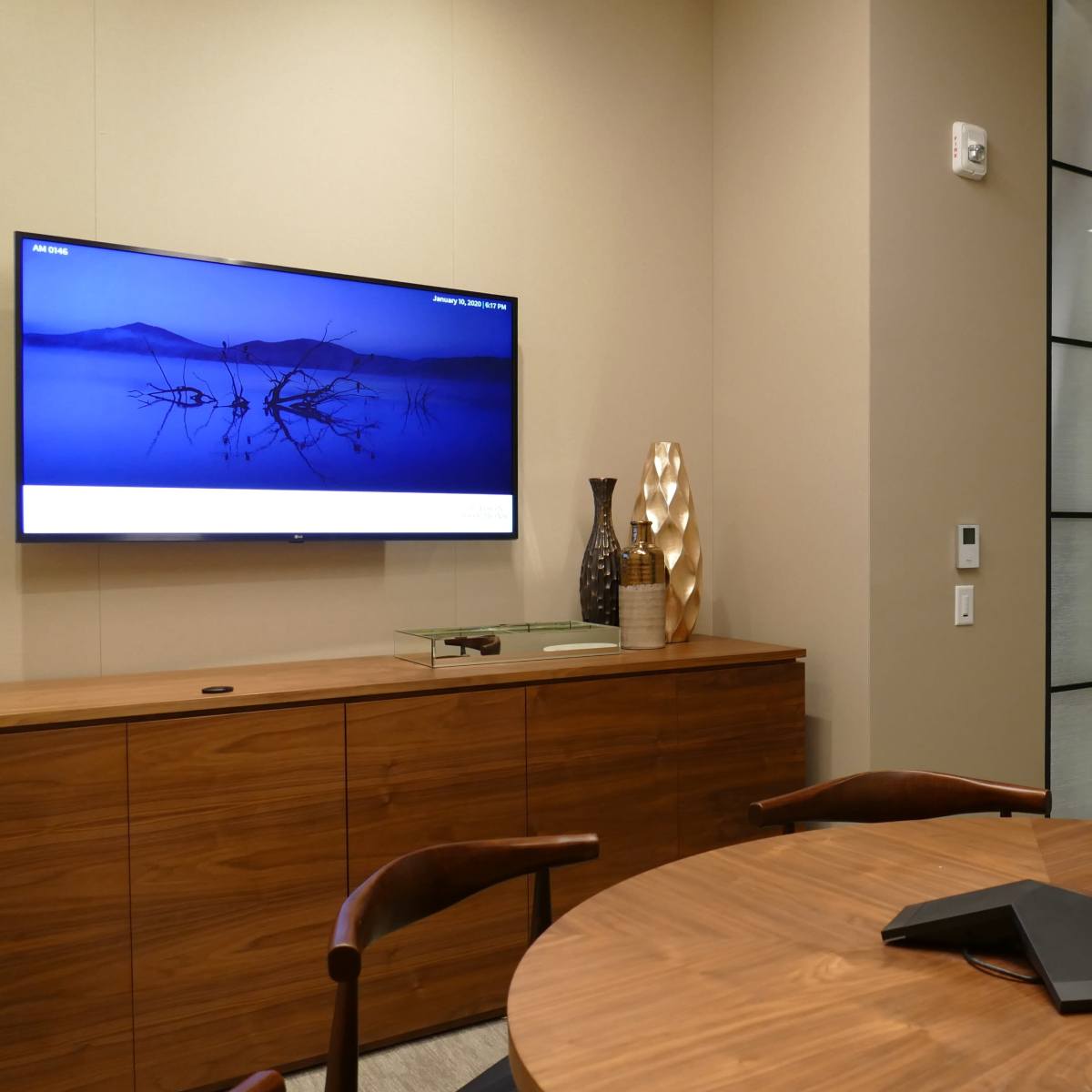 A modern conference room with a round table, a wall-mounted television displaying a blue image, and glass-paneled doors.