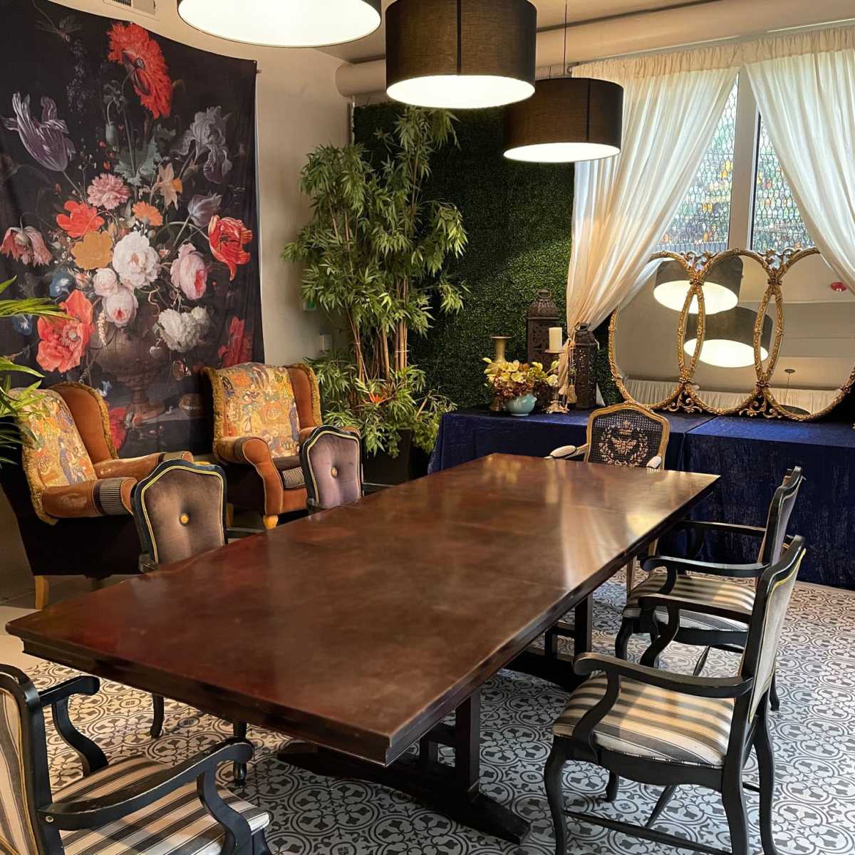A long wooden dining table is surrounded by several chairs in a room decorated with floral artwork, plants, and elegant lighting fixtures.