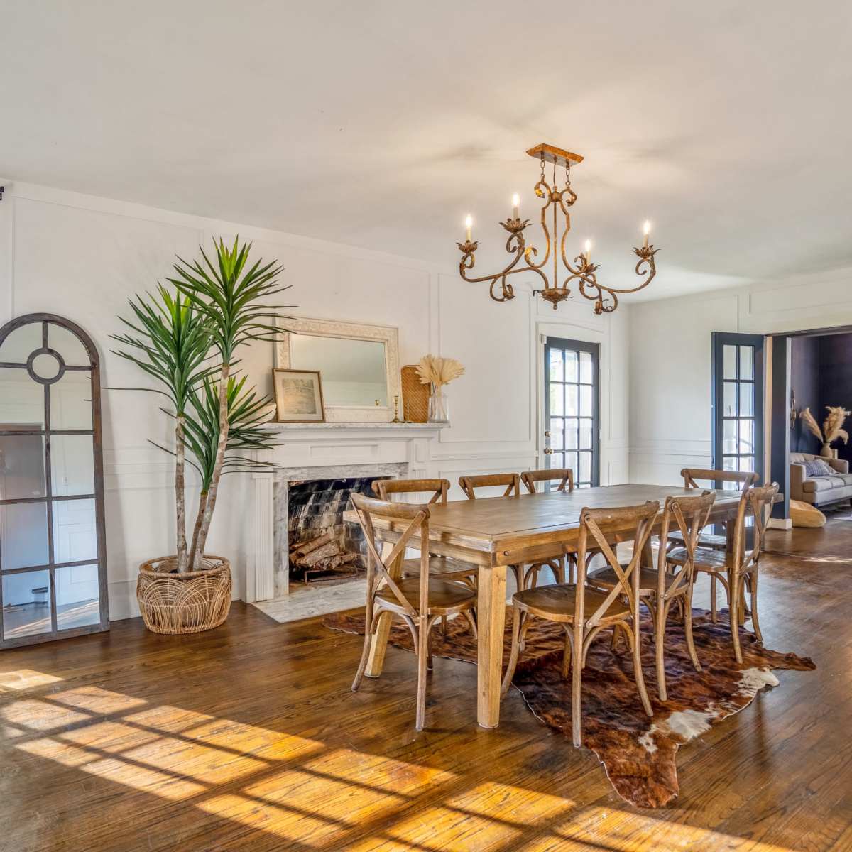 A spacious dining room with a wooden table surrounded by chairs, a chandelier overhead, and large windows allowing natural light to enter, alongside a fireplace and decorative elements.