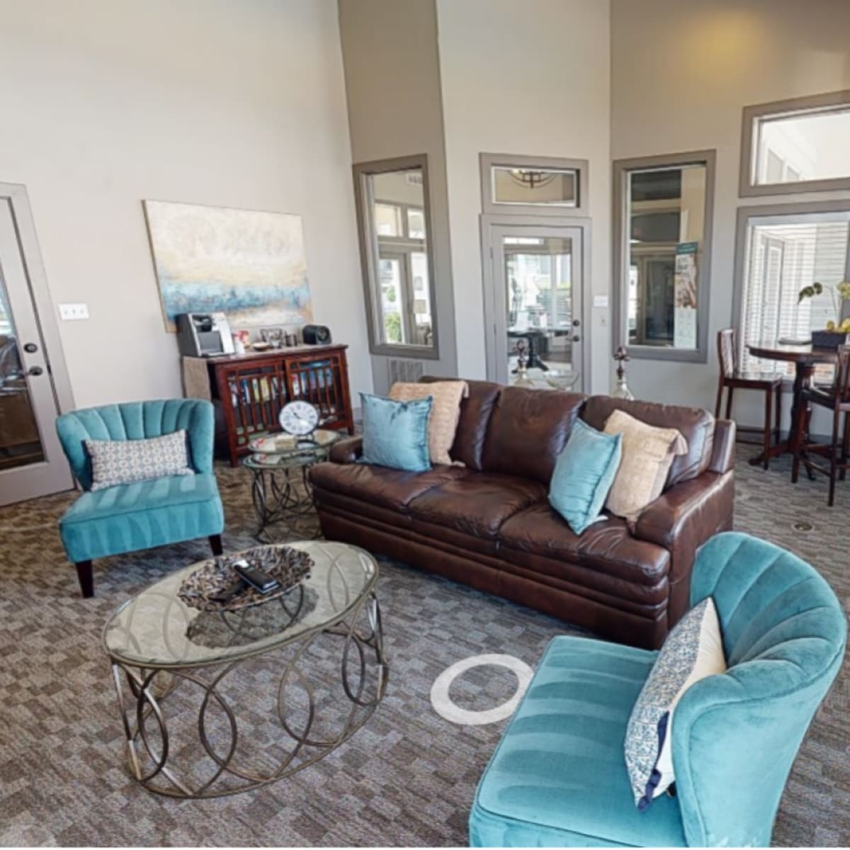 A modern lounge area featuring a brown leather sofa, turquoise armchairs, a glass coffee table, and a small dining table with chairs.
