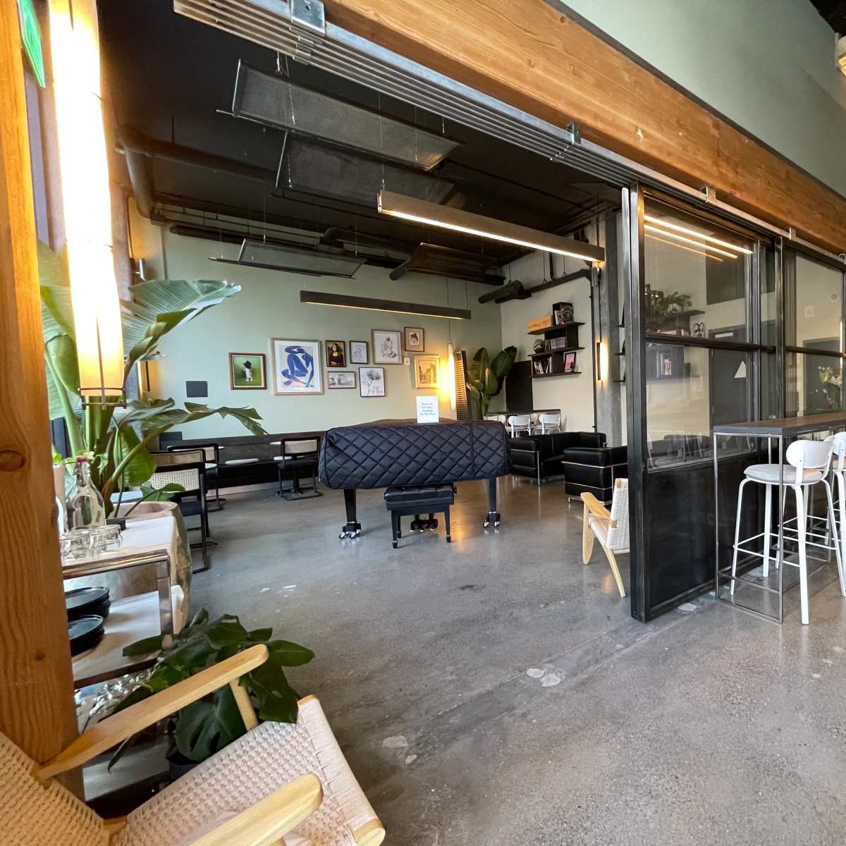 A modern interior of a cafe featuring a mix of seating areas, including a piano, bar stools, and plants.