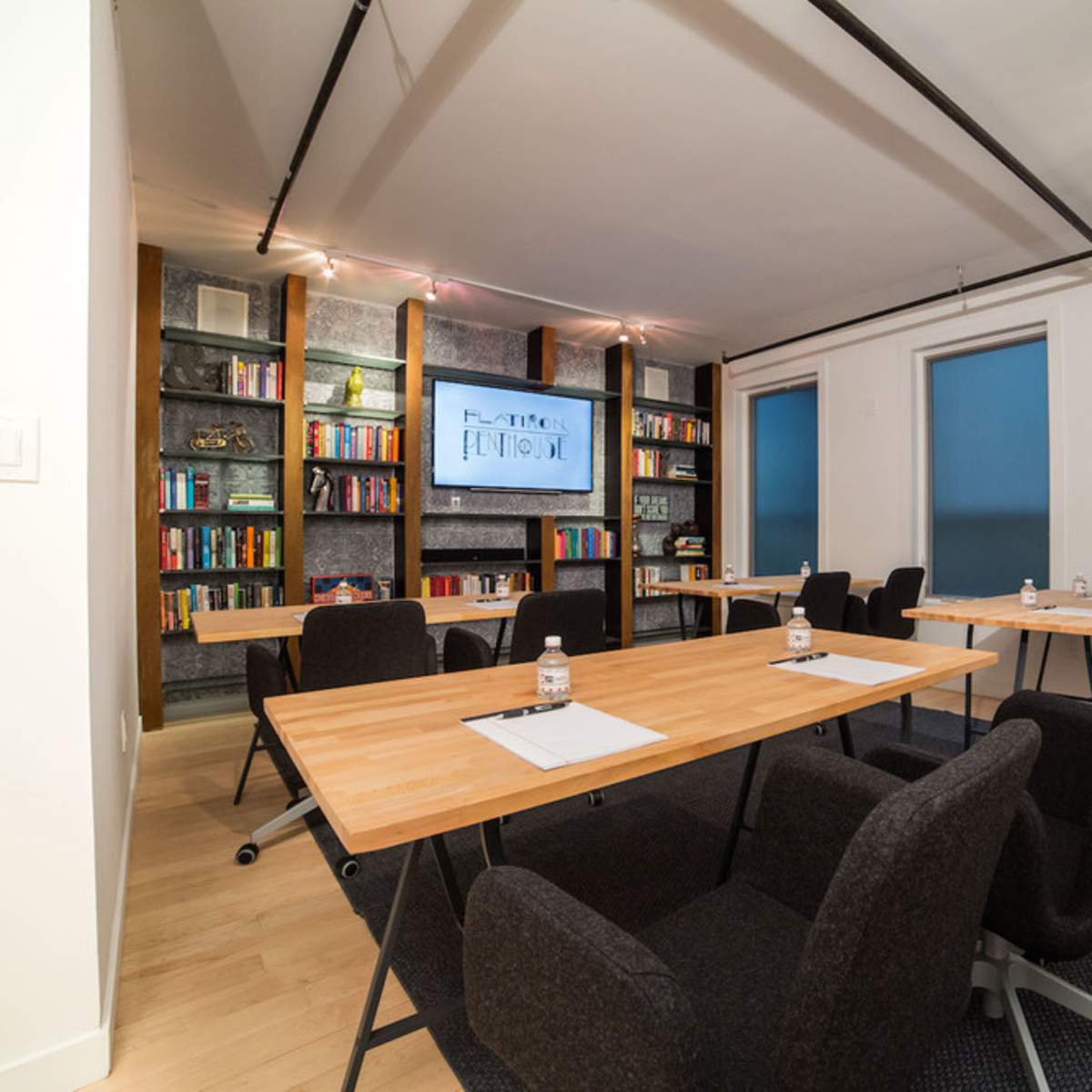 A meeting room features wooden tables arranged with chairs, a large screen on the wall, and bookshelves filled with books.