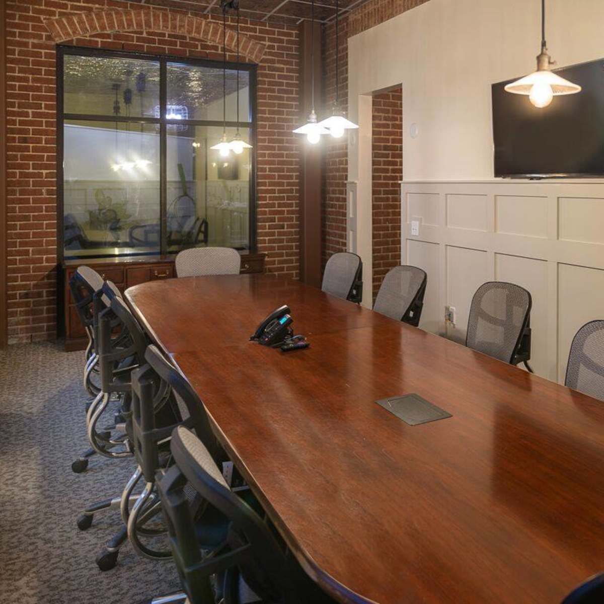 A long wooden conference table is surrounded by ergonomic chairs in a modern meeting room with a brick wall and a television mounted on one side.