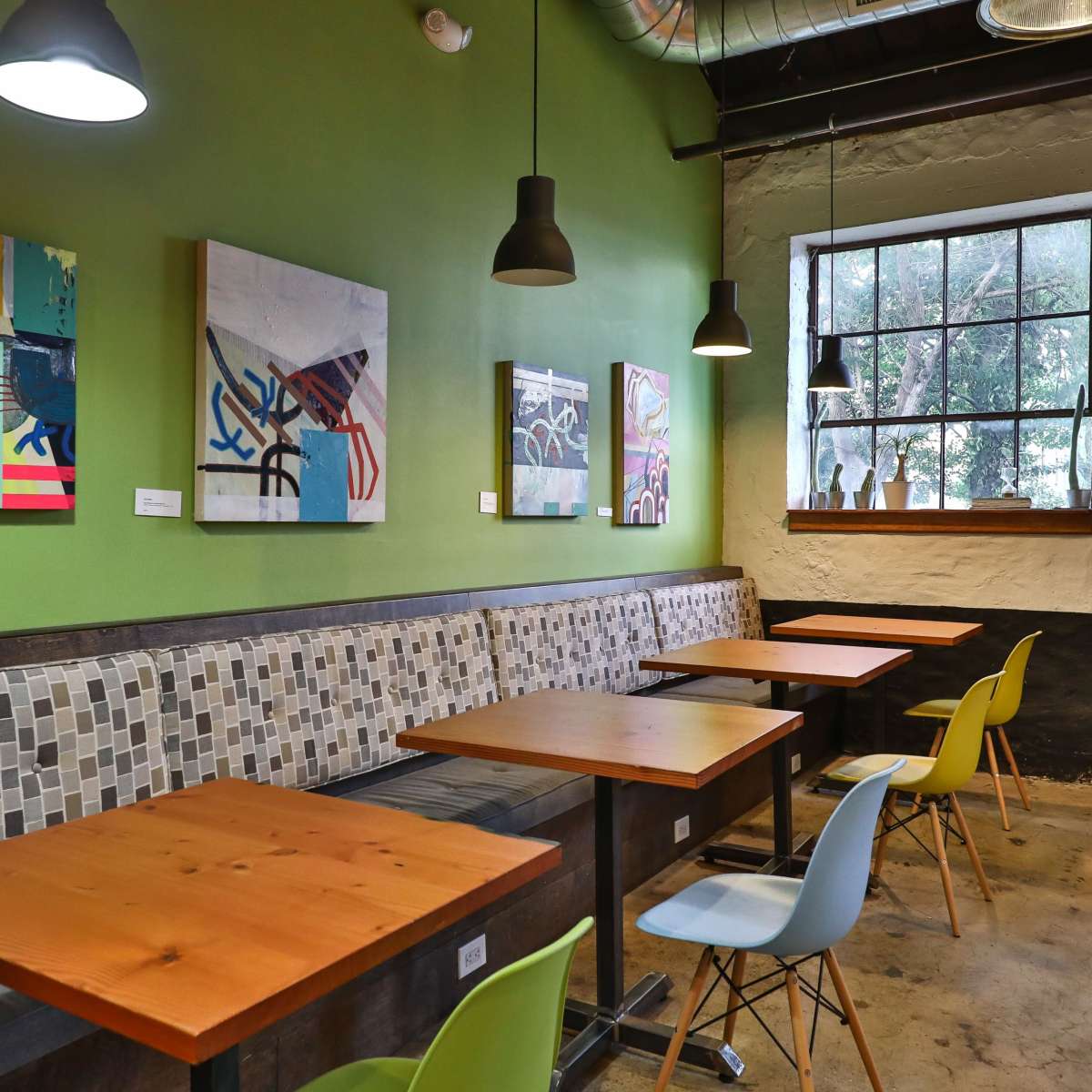 A cafe interior featuring a green wall, several pieces of abstract artwork, wooden tables, and a window with plants on the sill.