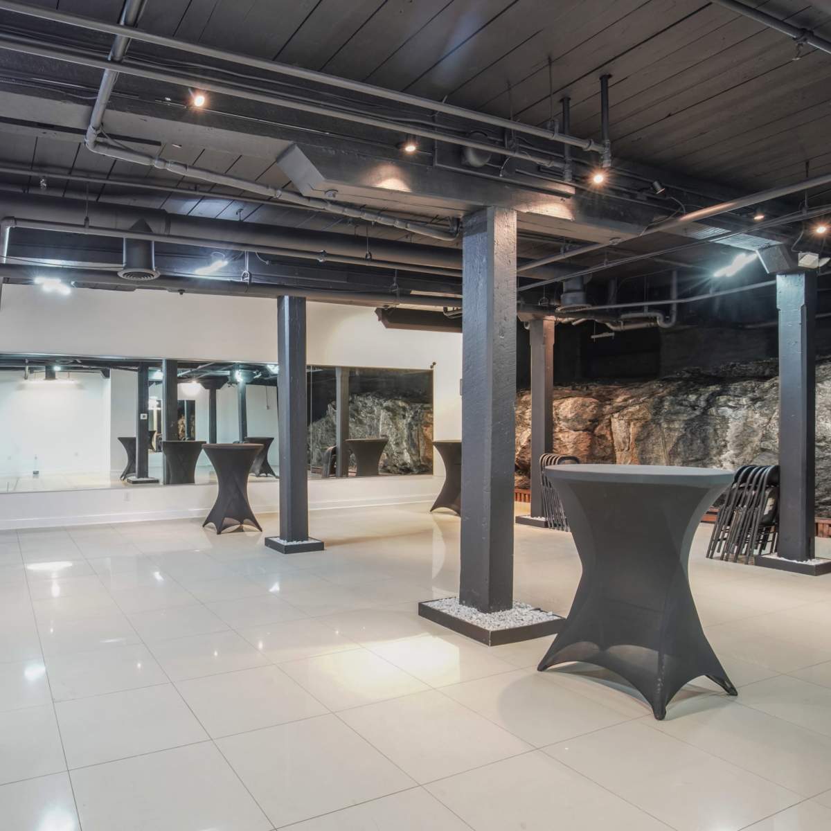 A spacious indoor area with black columns, tiled flooring, and multiple lighting fixtures, alongside mirrored walls and decorative features.