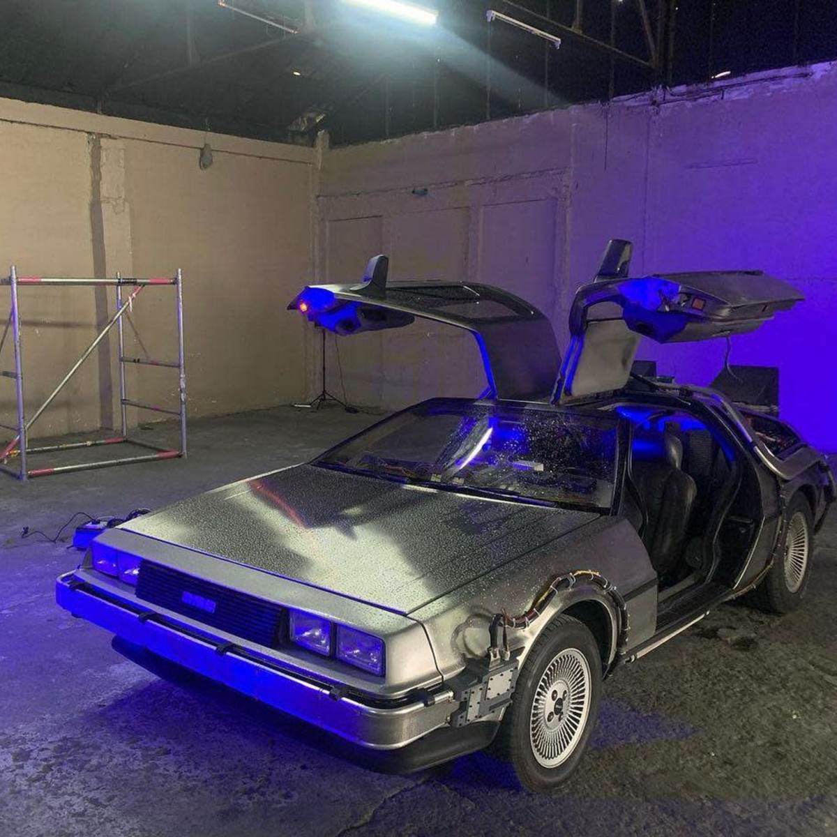 A DeLorean car with gull-wing doors is parked in an empty industrial space with a scaffold in the background and blue lighting.