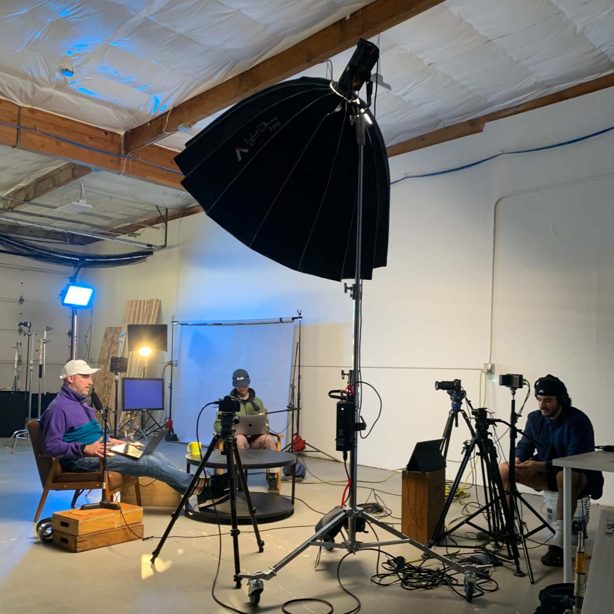 A group of three people is seated in a dimly lit studio with professional lighting and camera equipment set up for a video recording.