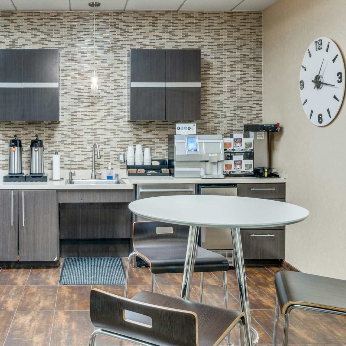 A modern kitchen area with a refrigerator, microwave, coffee dispensers, and a round dining table.