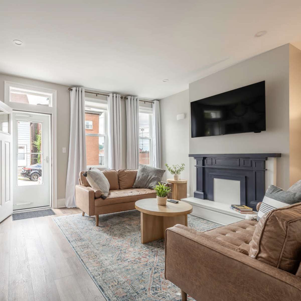 A modern living room featuring beige sofas, a round wooden coffee table, a flat-screen TV mounted on the wall, and large windows with sheer curtains.