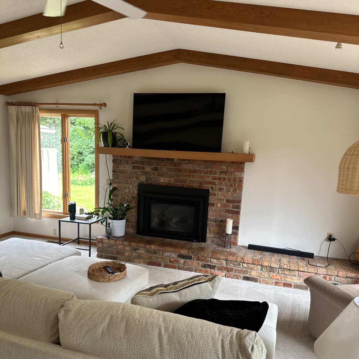 The living room features a brick fireplace with a mounted television above it, a large sectional sofa, and large windows that overlook a green outdoor view.