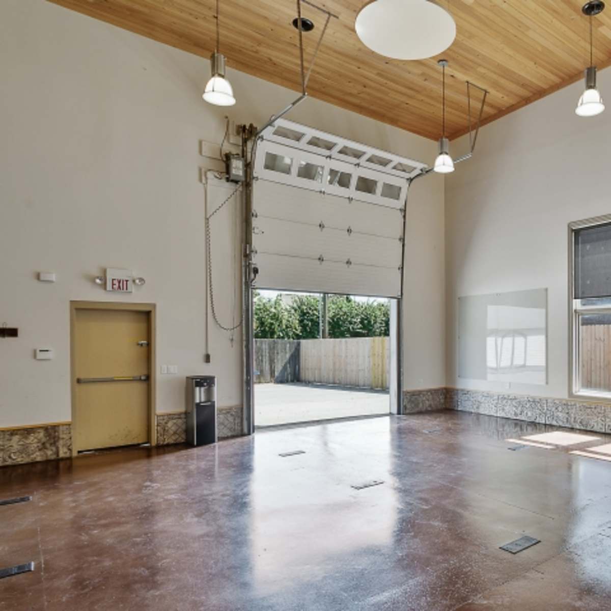 An empty interior space with a high ceiling, a large overhead door, and multiple windows letting in natural light.