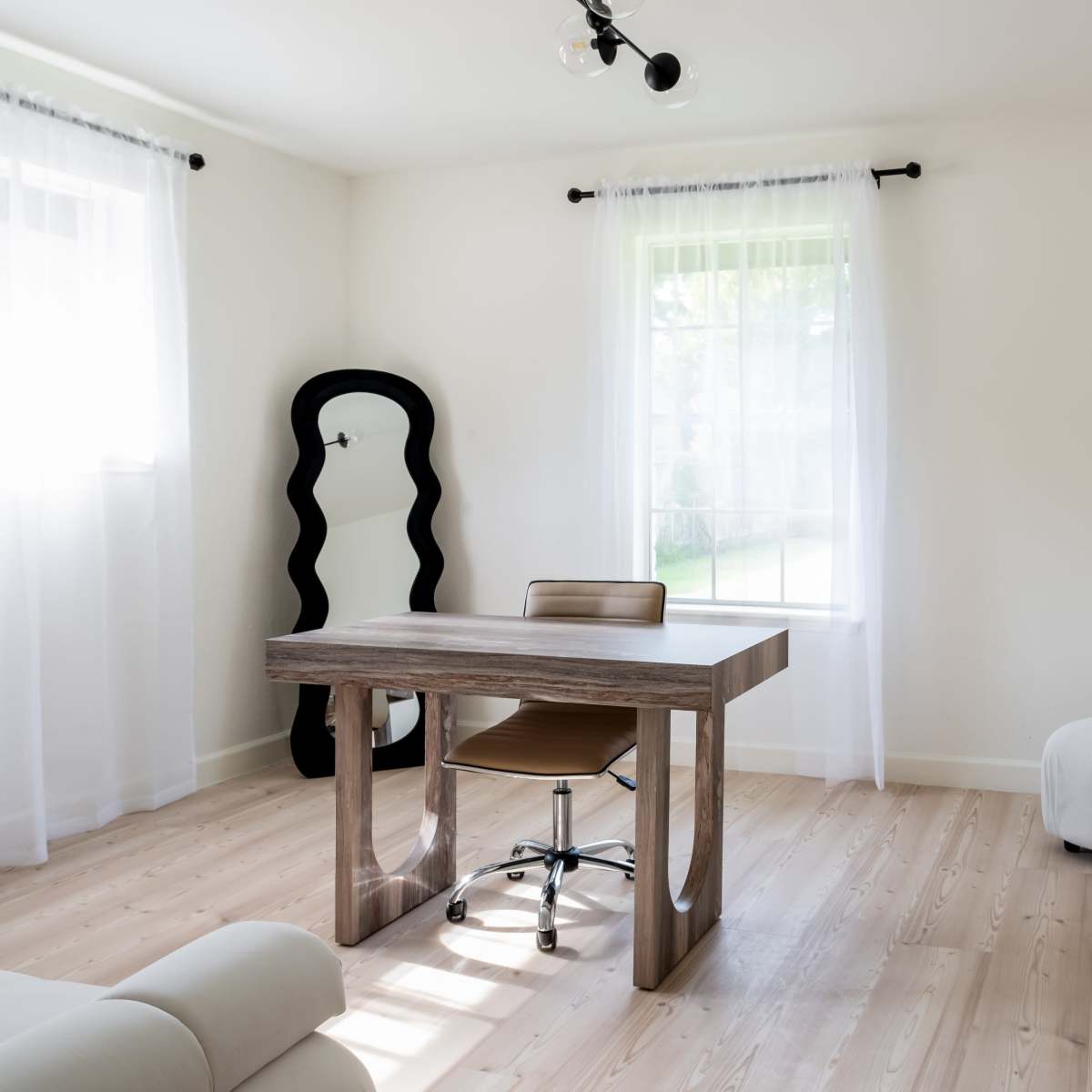 A minimalist room featuring a wooden desk with a chair, a soft couch, and large windows with sheer curtains.