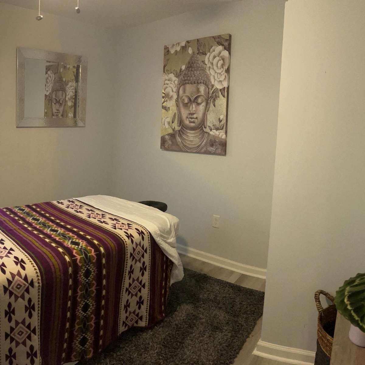The room features a massage table covered with a patterned blanket, a large Buddha artwork on the wall, and a mirror reflecting the space.